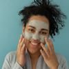 Positive ethnic female applying skincare clay mask for purifying pores and looking at camera against blue background