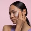 Young woman applying face cream with pink background, studio setting